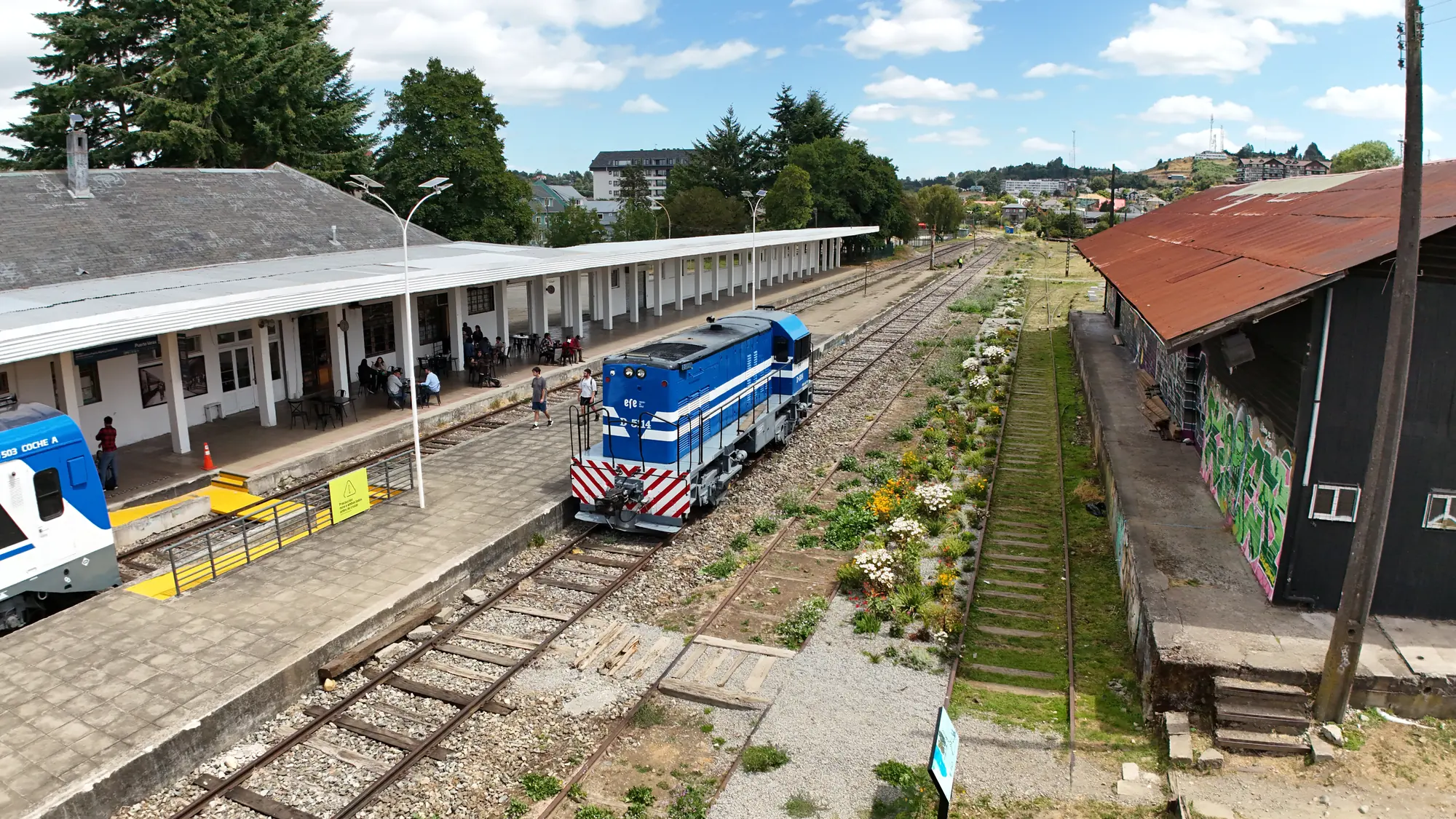 Locomotora azul entre el andén de la estación y el galpón, con las vías en perspectiva