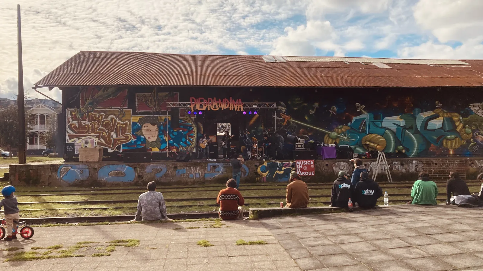 Banda tocando en un escenario frente al galpón, con público sentado en el andén